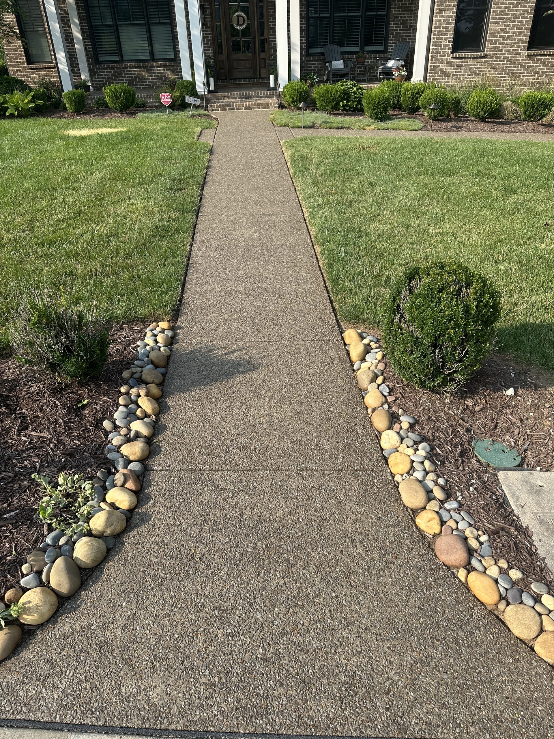 IMG_6155 Concrete walkway with stone borders leads to brick house, green lawn, shrubs Cody X Pressure Washing