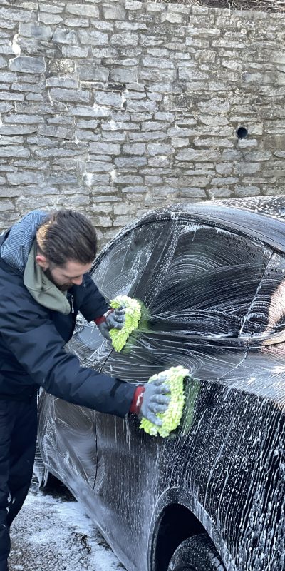 Person in black jacket washes black car with green mitts near stone wall. Cody-X Pressure Washing