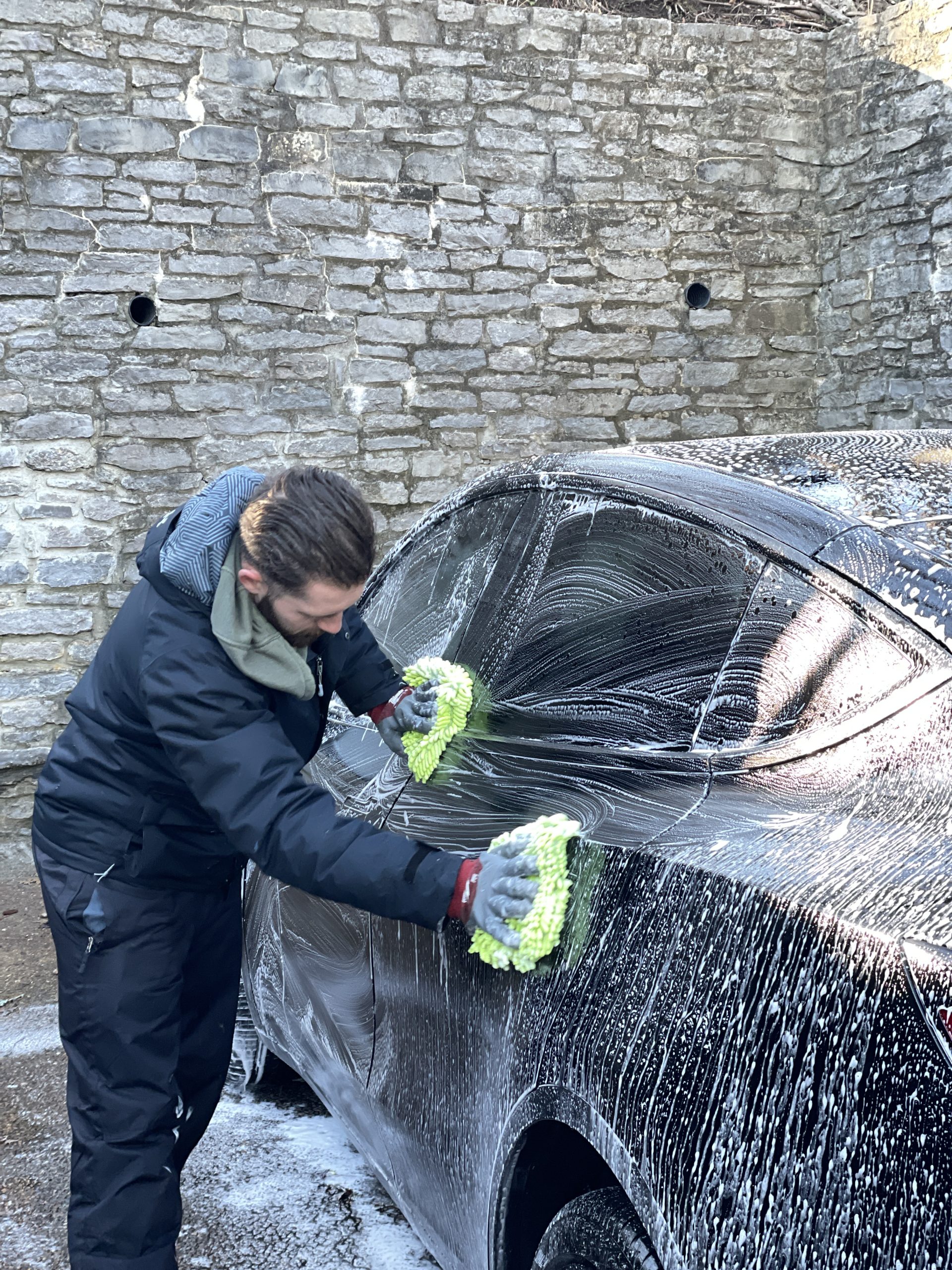 Photo Feb 01 2024, 8 45 57 AM Person in black jacket washes black car with green mitts near stone wall Cody X Pressure Washing