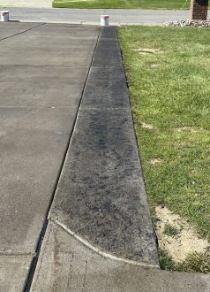 Photo May 25 2023, 4 35 33 PM A stained concrete curb separates pavement from grass; two buckets sit street-side. Cody-X Pressure Washing