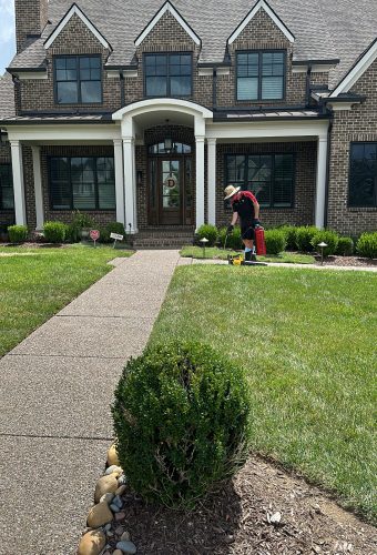 Photo Aug 02 2024, 2 07 47 PM Person with hat and backpack sprays lawn at large brick house; Cody-X service. Cody-X Pressure Washing