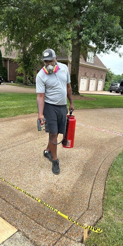 826AF884-4448-4C5C-AFB5-E74F428AEE44 Man in gear sprays driveway near caution tape with house and trees behind. Cody-X Pressure Washing