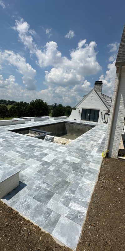 Partially built pool with stone tiles beside a white house under clouds. Cody-X Pressure Washing