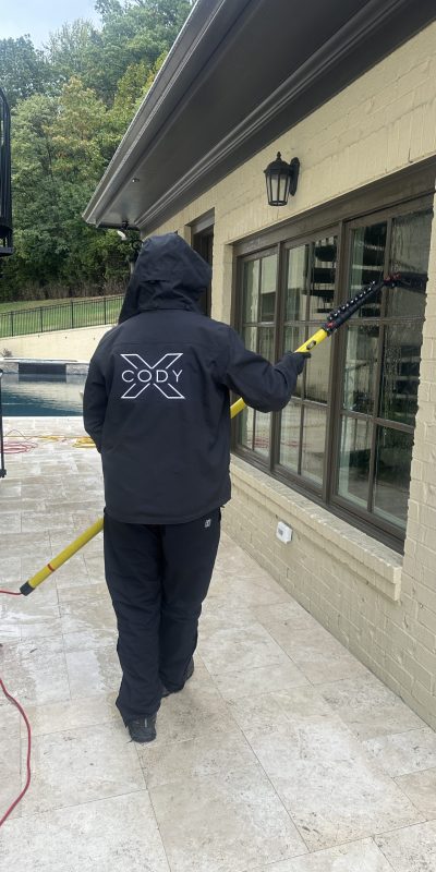 Person in "CODY" jacket cleans building windows; red cord on patio. Cody-X Pressure Washing