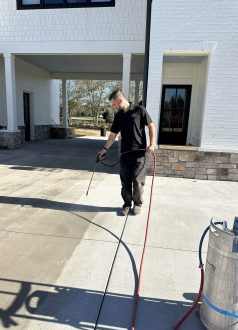 IMG_1149 A man pressure washes a concrete driveway outside a white brick house. Cody-X Pressure Washing