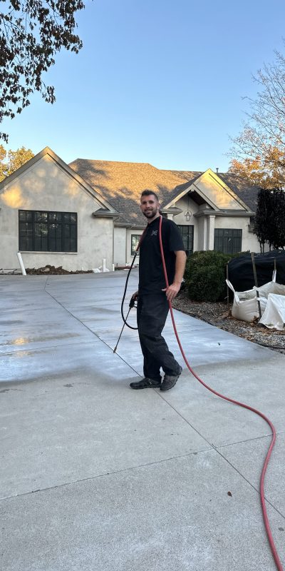 A man with a red hose stands on a new driveway, tools and materials nearby. Cody-X pressure washing and sealing service. Cody-X Pressure Washing